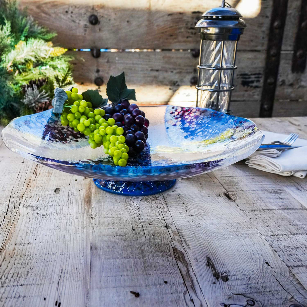 A beautiful purple, blue and clear serving pedestal bowl with grapes set on it.