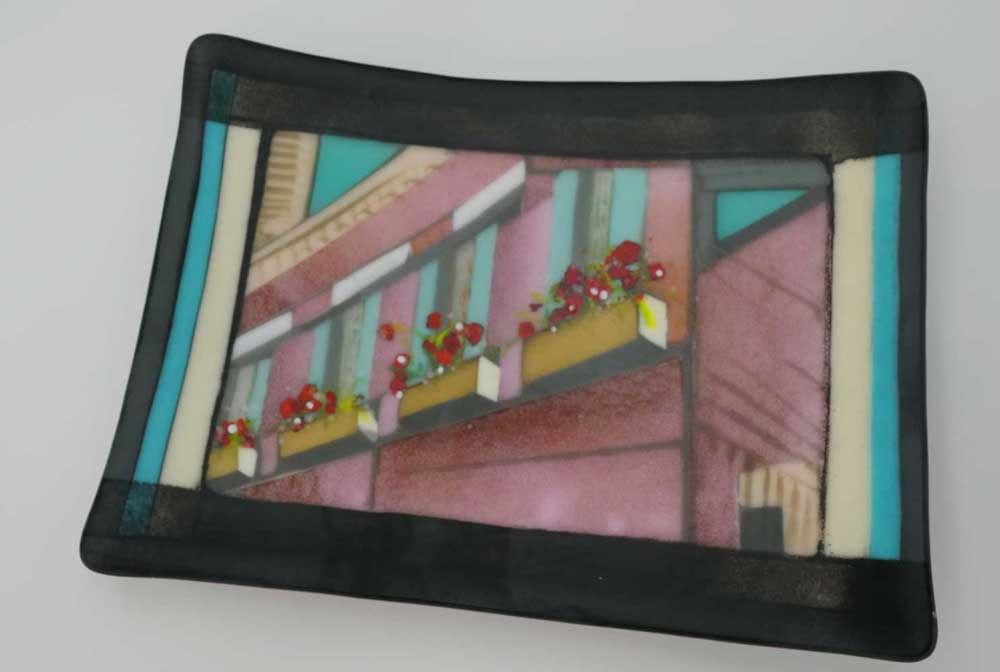 An Art Glass platter that is an abstract building scene of a Venice street. Dominate colors are pink, light blue, yellow and grey. 