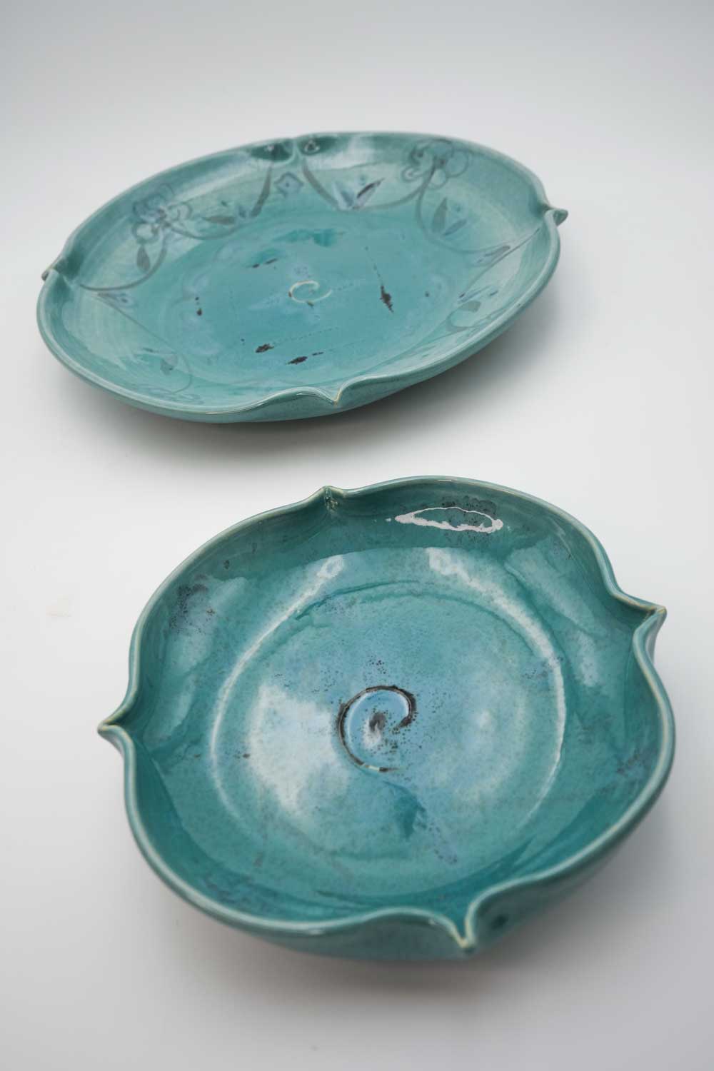 Two handmade ceramic wide and shallow turquoise bowls with fluted edges. Decorated with painted grey floral design. 
