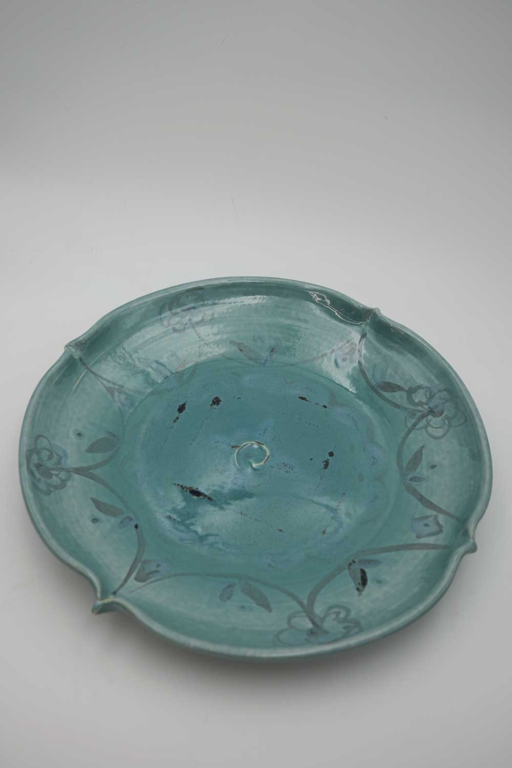 A  handmade ceramic wide and shallow turquoise bowl with fluted edges. Decorated with painted grey floral design. 